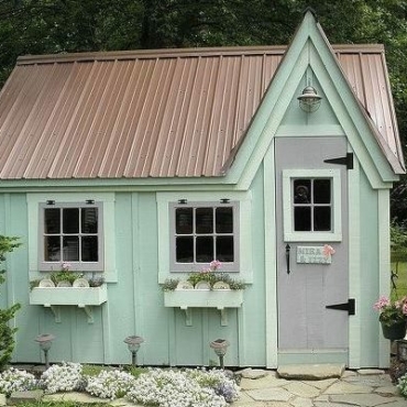 The exterior of a quaint garden shed, painted pale green, with a pitched roof and window boxes filled with flowers
