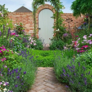 a small garden with overflowing flower beds and a brick path leading to a pretty gate at the end