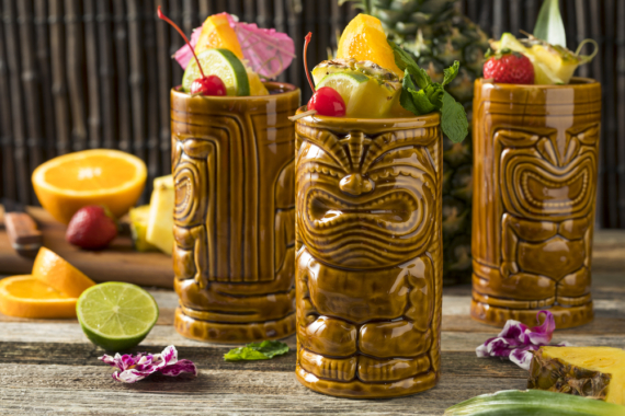 three novelty ceramic tiki mugs shaped like wood carvings are filled with exotic fruit drinks